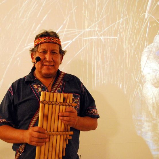 Ein Mann mit Mikrofon und Stirnband steht und hält eine Panflöte vor einer weißen Wand mit Grasprojektionen.