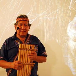 Ein Mann mit Mikrofon und Stirnband steht und hält eine Panflöte vor einer weißen Wand mit Grasprojektionen.