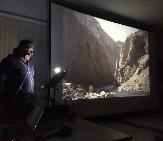 Ein Mann steht in einem schwach beleuchteten Raum, vor einer Projektionsleinwand, die eine Berglandschaft zeigt. Ein Laptop und eine Lampe sind auf einem Tisch vor ihm platziert.