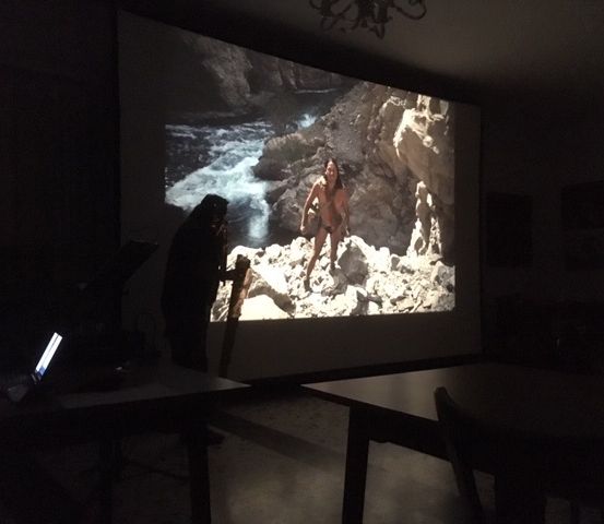 Eine Person steht in einem schwach beleuchteten Raum und beobachtet eine große Leinwand, auf der eine Szene eines Mannes zu sehen ist, der auf Felsen in der Nähe eines Wasserfalls steht. Eine weitere Person sitzt in der Nähe.