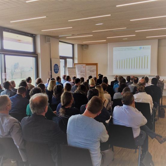 Ein Konferenzraum mit Menschen in Stühlen, die einer Präsentation zuhören. Eine Projektionsleinwand zeigt ein Diagramm. Ein Whiteboard steht neben der Leinwand. Es gibt Glasfenster mit Blick auf Bäume im Freien.