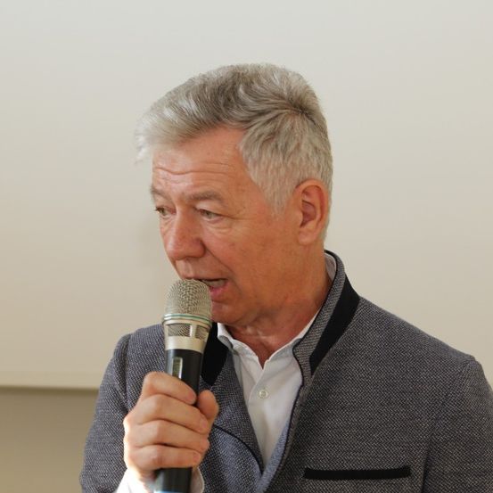 Ein älterer Mann mit grauem Haar hält ein Mikrofon und scheint bei einer Veranstaltung zu sprechen. Er trägt eine graue Jacke und ein weißes Hemd.