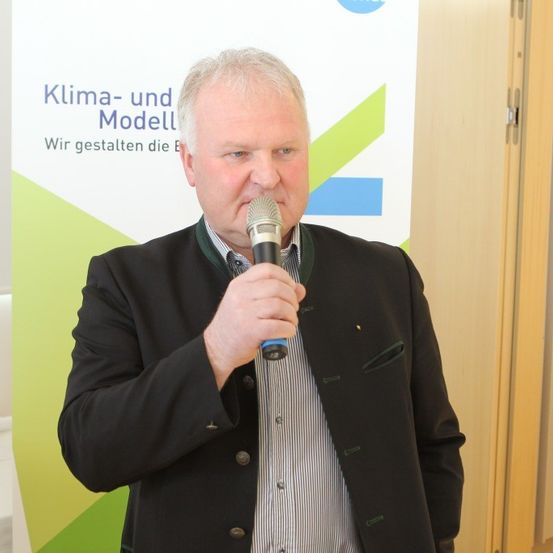 Ein Mann in einem Anzug hält ein Mikrofon und spricht auf einer Konferenz. Hinter ihm steht ein Banner mit der Aufschrift 'Klima- und Modell'.
