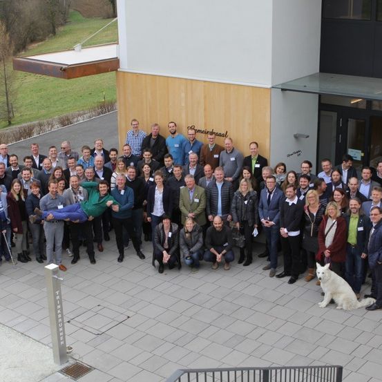 Eine Gruppe von Menschen in formeller Kleidung versammelt sich vor einem Gebäude mit einem Grasfeld im Hintergrund. Ein Hund steht im Vordergrund.