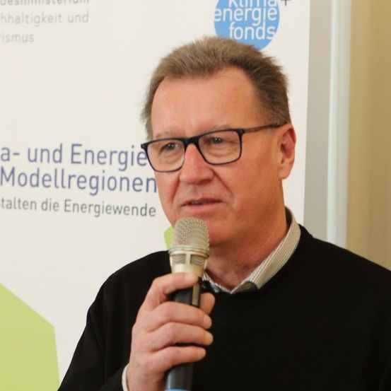 Ein Mann mit Brille und schwarzem Pullover steht vor einem Mikrofon und hält vermutlich eine Rede. Hinter ihm ist eine Hintergrundkulisse mit grünem und weißem Text, der 'klima- und Energie Modellregionen' lautet.