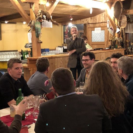 Ein Mann steht und spricht in ein Mikrofon in einem Restaurant, während die Leute an Tischen sitzen. Ein hölzerner Pfeiler hält einen hängenden Affen.