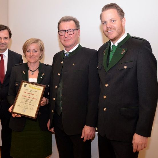 Eine Gruppe von vier Personen steht zusammen und hält eine Plakette, zwei Männer und zwei Frauen, alle in formeller Kleidung. Die Frau auf der linken Seite hält die Plakette.