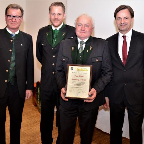 Vier Männer stehen zusammen, einer in der Mitte hält ein gerahmtes Zertifikat. Der Mann in der Mitte trägt eine Medaille um den Hals. Hinter ihm stehen zwei Männer in formeller Kleidung und einer in einem grünen Weste. Sie alle lächeln.