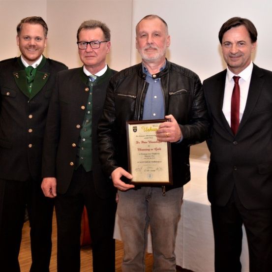 Vier Männer stehen zusammen, einer hält eine Urkunde. Sie sind in formeller Kleidung gekleidet, wahrscheinlich für eine Preisverleihung. Hinter ihnen ist eine weiße Wand und eine weiße Tischdecke.