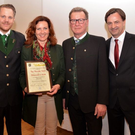 Eine Frau in einer grünen Jacke hält eine gerahmte Urkunde, während vier Männer hinter ihr stehen. Sie alle schauen in die Kamera und lächeln. Der Raum hat eine weiße Wand und einen Holzboden.