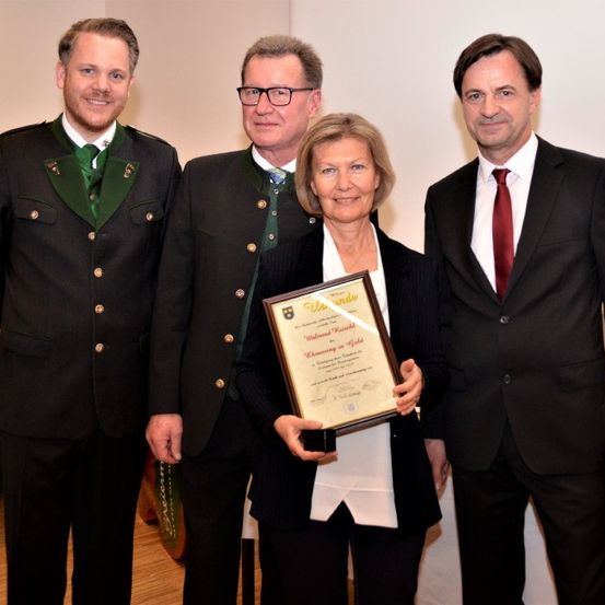 Vier Personen stehen zusammen, zwei Männer in traditionellen Uniformen und zwei in Anzügen, lächeln in die Kamera. Eine Frau hält ein gerahmtes Zertifikat in den Händen.