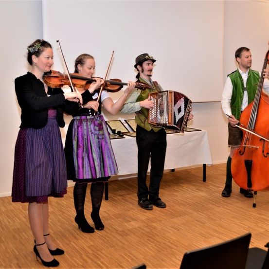 Vier Personen spielen in einem Raum, zwei mit Violinen und zwei mit größeren Instrumenten, möglicherweise Cello und Akkordeon. Der Raum hat Holzboden und eine weiße Rückwand.