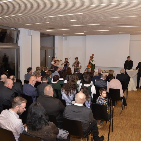 Ein Publikum in formeller Kleidung versammelt sich in einem Raum mit Holzböden und beobachtet eine Aufführung eines Streichquartetts und eines Akkordeonspielers. Zwei Männer stehen an einem Tisch auf der rechten Seite.