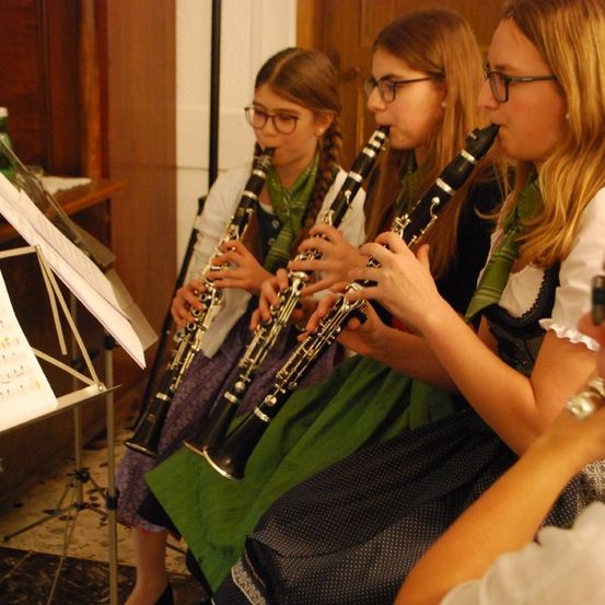 Vier Mädchen in traditioneller Kleidung spielen Klarinetten in einem Raum mit Notenständern, Flaschen auf einem Tisch und einem Teppichboden.