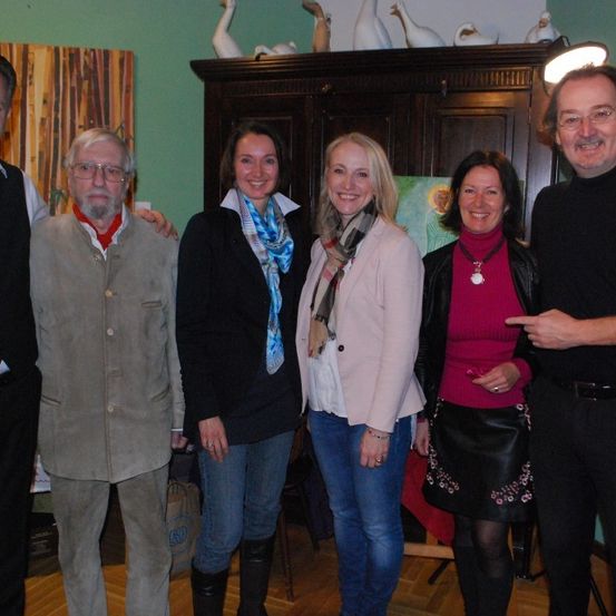 Sechs Personen posieren für ein Foto im Innenbereich. Sie sind in lässiger Winterkleidung gekleidet. Hinter ihnen steht ein hölzerner Schrank, der mit dekorativen Gegenständen verziert ist.