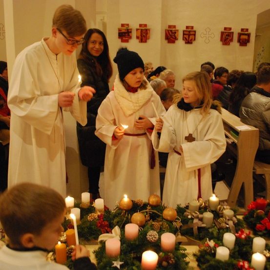 Ein Priester in einer weißen Robe zündet mit zwei jungen Mädchen Kerzen während einer Zeremonie in einer Kirche an. Die Mädchen halten ebenfalls Kerzen. Andere Menschen sitzen auf Bänken hinter ihnen. Der Altar ist mit Kränzen, Kerzen und Ornamenten geschmückt.