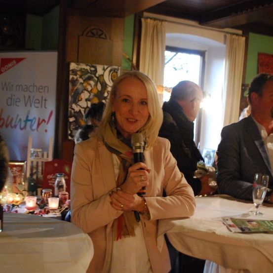 Eine Frau hält ein Mikrofon und lächelt, steht vor einem Tisch mit Kerzen. Dahinter sitzen zwei Männer, einer mit einem Glas Wein. Ein Banner lautet 'Wir machen die Welt besser!' Ein Fenster mit Vorhängen im Hintergrund.