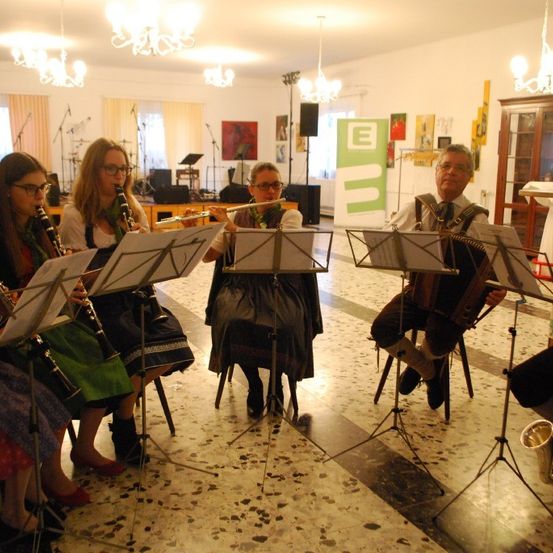 Eine Gruppe von Musikern spielt Klarinetten, Flöten und ein Akkordeon, mit Notenständern vor ihnen. Die Umgebung ist drinnen mit Kronleuchtern und Gemälden.