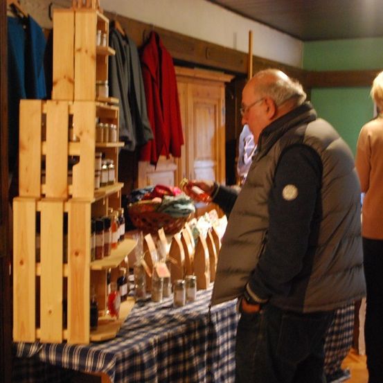 Ein älterer Mann mit Brille untersucht Gegenstände auf einem Tisch, mit einem Holzregal voller Gewürze hinter ihm. Eine Frau steht neben ihm.