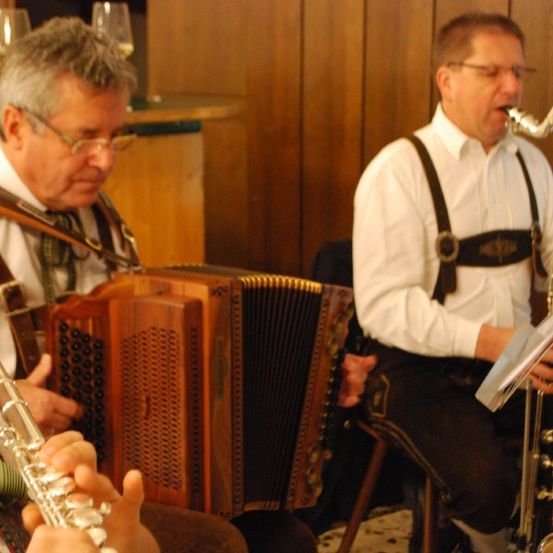 Zwei Männer in traditioneller Kleidung spielen Musikinstrumente in einem Raum. Einer spielt die Flöte und der andere das Saxophon.