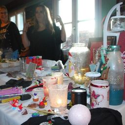 Zwei Frauen stehen hinter einem Tisch mit verschiedenen Bastelutensilien. Es gibt Gläser, Kerzen, Cupcakes und andere Gegenstände auf dem Tisch. Eine Frau lächelt und hebt ihre Hand.