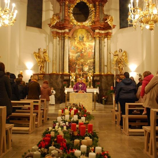 Ein Kircheninneres mit einem Priester am Altar. Kerzen säumen den Weg zum Altar, und Menschen sitzen auf Bänken. Es gibt goldene Statuen und Kronleuchter.