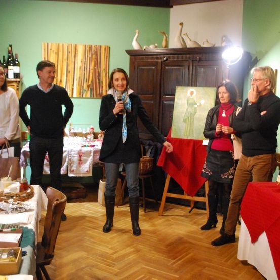 Eine Gruppe von Menschen in einem Raum, eine Frau mit einem Mikrofon steht in der Mitte. Ein Gemälde auf einem hölzernen Staffelei und Tische mit verschiedenen Gegenständen sind in der Nähe. Ein hölzerner Schrank mit Weinflaschen und dekorativen Vögeln steht an der Wand.