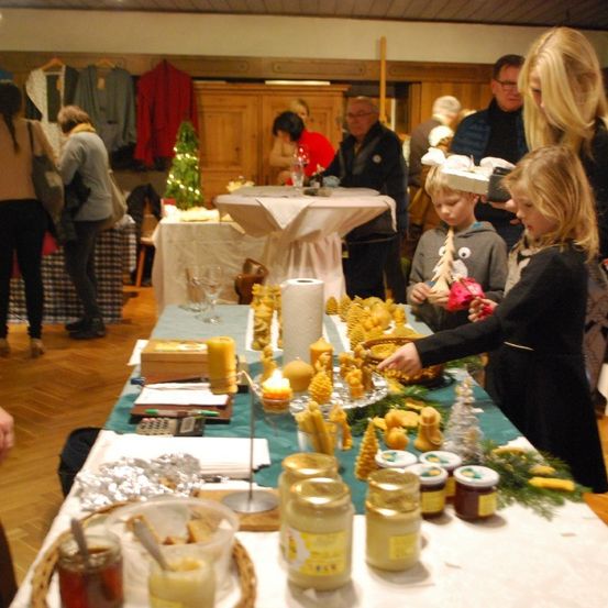 Eine Gruppe von Menschen versammelt sich in einem Raum mit einem langen Tisch, der mit Kerzen, Honiggläsern und anderen Gegenständen gefüllt ist. Einige betrachten die Gegenstände, während andere sich unterhalten. Der Raum hat Holzböden und -wände.