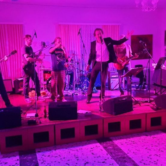 Eine Band spielt auf einer Bühne mit lila Beleuchtung. Ein Mann singt in ein Mikrofon, während andere Gitarre und Schlagzeug spielen.