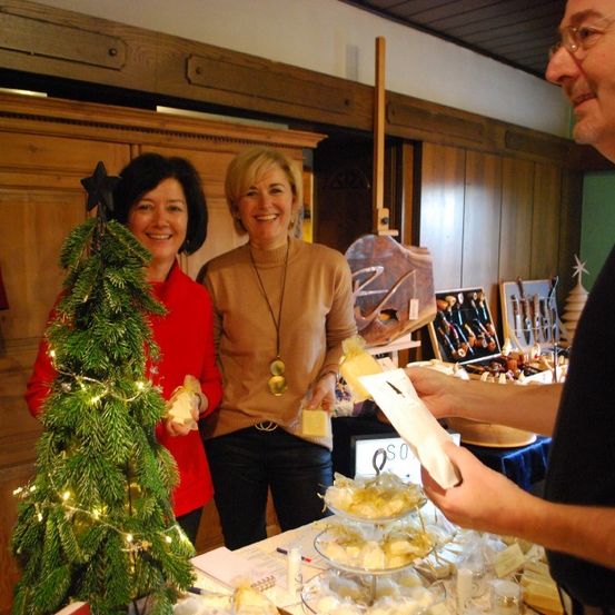 Zwei Frauen lächeln, eine hält einen kleinen Weihnachtsbaum, die andere eine Tasche. Ein Mann neben ihnen hält ein Papier. Vor ihnen steht ein Tisch mit Essen und einer digitalen Anzeige.