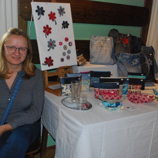 Eine Frau mit Brille sitzt neben einem Tisch mit verschiedenen Gegenständen, darunter Taschen, Beutel und eine weiße Leinwand mit Blumendesigns.