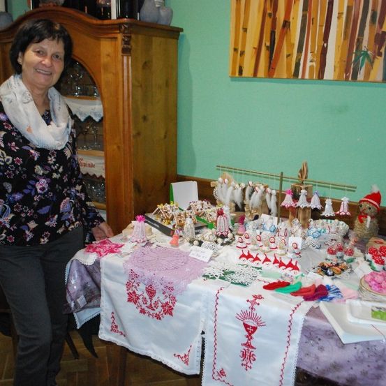 Eine Frau steht neben einem Tisch, der mit verschiedenen dekorativen Gegenständen und Kunsthandwerken gefüllt ist, darunter Spitze, Ornamente und kleine Skulpturen. Hinter ihr ist ein hölzerner Schrank und eine Wand mit einem Bambus-Gemälde zu sehen.