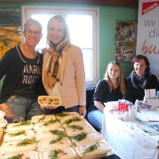 Vier Frauen stehen an einem Tisch mit Essen. Eine hält eine Platte mit Essen. Eine andere hat eine Brille und ein schwarzes Shirt. Der Tisch ist mit einem weißen Tuch bedeckt.