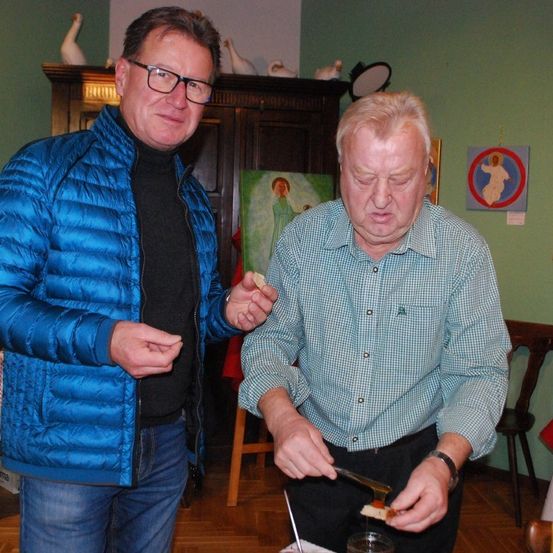 Zwei Männer sind in einem Raum. Einer hält Essen und schaut es sich an. Der andere schmiert Honig auf ein Stück Brot. Es hängen Gemälde an der Wand.