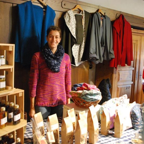 Eine Frau steht vor einem Tisch mit mehreren Gläsern und Tüten. Hinter ihr hängen verschiedene Kleidungsstücke an der Wand. Links von ihr steht ein Regal mit Flaschen und Gläsern und rechts von ihr ein Holzschrank.