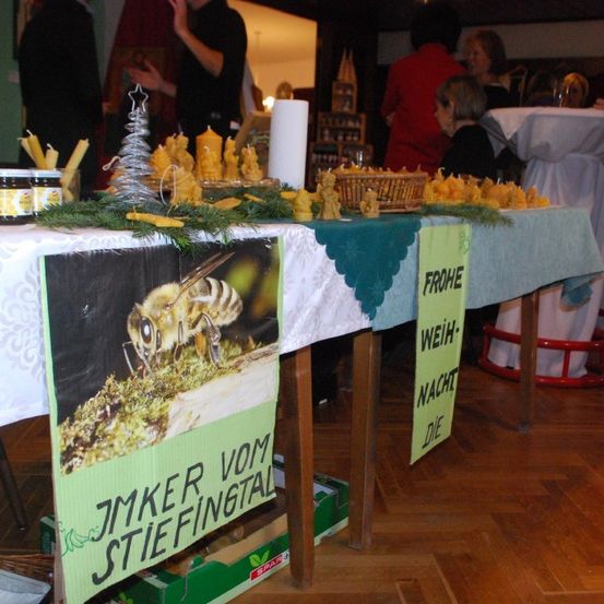 Ein Tisch mit einem Banner einer Biene und einem Plakat mit der Aufschrift 'Jmker vom Stiftungsfal'. Verschiedene Produkte, möglicherweise Honig, sind auf dem Tisch ausgestellt. Ein gelbes Schild steht 'VON WEIHNACHTSNACHT BIS DIE'.