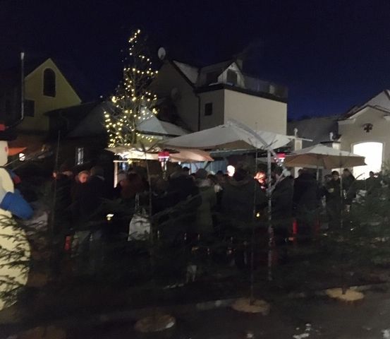 Eine Menschenmenge versammelt sich am Abend um einen geschmückten Weihnachtsbaum, mit mehreren Schirmen und einem großen Schneemann in der Nähe.