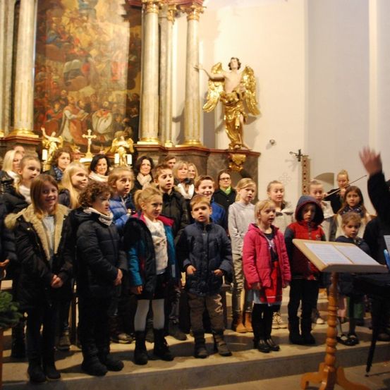 Eine Gruppe von Kindern in Wintermänteln steht auf den Altarstufen einer Kirche und singt, während ein Erwachsener Geige spielt.