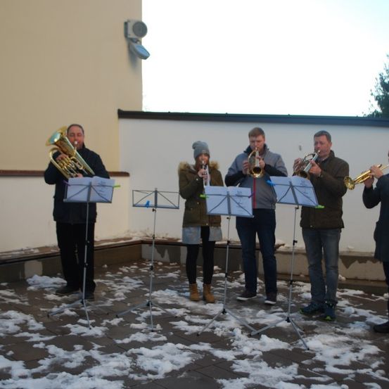 Sechs Personen stehen auf einer verschneiten Oberfläche und spielen Trompeten. Alle sind warm gekleidet, mit Notenständern vor ihnen. Im Hintergrund befindet sich eine Wand mit einem Fenster und einem Baum.