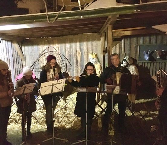 Eine Gruppe von Musikern spielt Instrumente in einer Scheune, bei schwachem Licht. Sie sind warm gekleidet, und die Umgebung deutet auf eine festliche Atmosphäre hin.