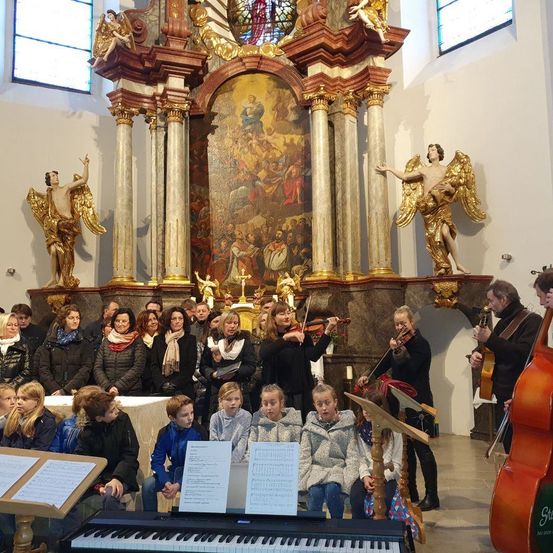 Ein Chor aus Kindern und Erwachsenen singt in einer Kirche mit Musikern, die Violinen und ein Cello spielen, vor einem Klavier. Ein Gemälde ist auf dem Altar.