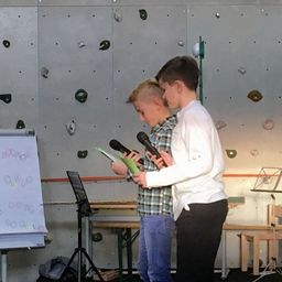 Zwei Jungen mit Mikrofonen stehen neben einer Tafel mit Musiknoten in einem Raum mit einer Kletterwand.
