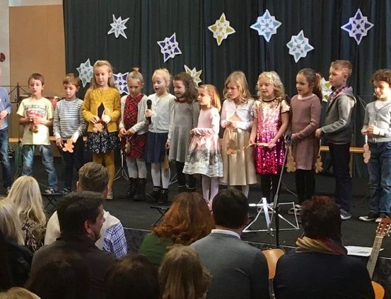 Eine Gruppe von Kindern tritt auf einer Bühne auf, hält Instrumente und trägt festliche Kleidung. Hinter ihnen zeigt ein Hintergrund Schneeflocken, und ein Publikum schaut zu.