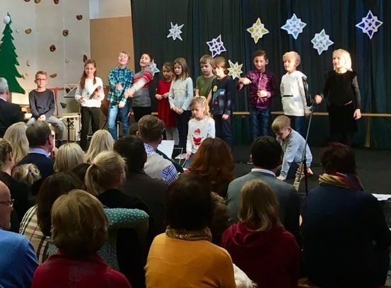 Eine Gruppe von Kindern in Winterkleidung steht auf der Bühne vor einem Publikum, geschmückt mit Weihnachtsdekorationen.