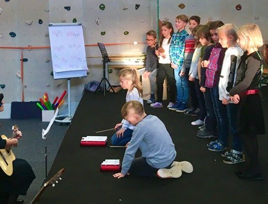 Eine Gruppe von Kindern versammelt sich in einem Musikraum, in dem eine Frau eine Gitarre spielt. Einige Kinder spielen Instrumente auf dem Boden.