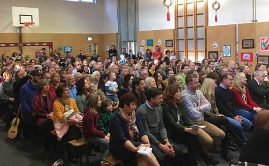 Ein vollbesetzter Raum mit einem Basketballkorb und einer Bühne, wo die Menschen auf Bänken sitzen, eine Person ein Buch hält und Kunst an der Wand hängt.