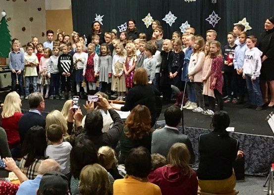 Eine Gruppe von Kindern steht mit Erwachsenen auf der Bühne, wahrscheinlich während einer Weihnachtsaufführung. Zuschauer sehen zu, einige machen Fotos. Ein Notenständer steht auf der Bühne.