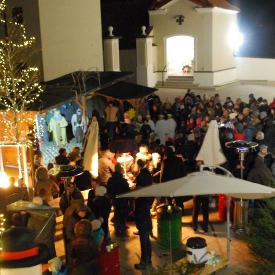 Eine Menschenmenge versammelt sich bei Nacht um eine Krippe, mit einer Kirche im Hintergrund, festliche Lichter erhellen die Szene, und Menschen stehen unter Regenschirmen und in der Nähe eines Feuerbeckens.