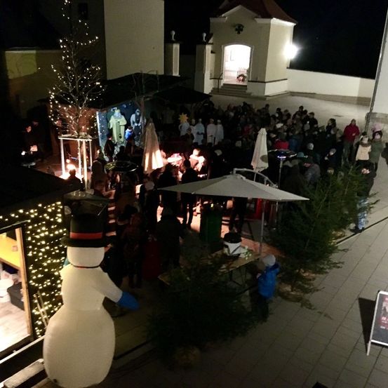 Eine Menschenmenge versammelt sich bei Nacht, umgeben von beleuchteten Bäumen und einem Schneemann. Einige stehen in der Nähe eines Tisches mit einem Schirm. Im Hintergrund sind eine Kirche und ein Gebäude zu sehen.