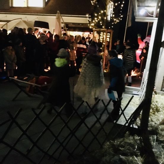 Eine Menschenmenge versammelt sich bei Nacht vor einem Gebäude. Einige tragen Winterkleidung. Es gibt Bäume und Lichter am Gebäude. Eine Person steht vor einem Feuer.